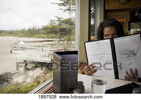 Stock Photo - A Woman Sitting In A Restaurant Peering Over The Menu Out The Window At The View; Tofino, British Columbia, Canada. Fotosearch