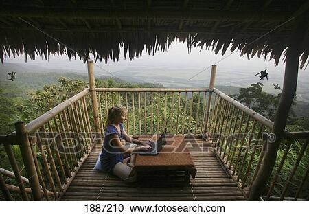 Stock Image - A Woman Sitting On The Floor Using A Wireless Laptop Computer To Get Email On The Balcony Of A Remote Bamboo Hut Overlooking Bangladesh. Fotosearch