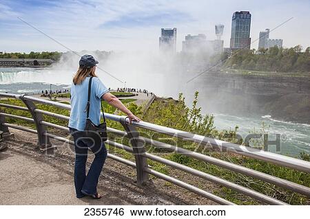 Stock Photograph - A Woman Stands At A Railing Overlooking Horseshoe Falls At Niagara Falls State Park, In New York State With The Town Of Niagara Falls, Ontario In The Background; Niagara Falls, New York, United States Of America. Fotosearch