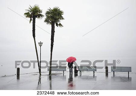 Stock Photo - A Woman Stands At The Water's Edge With A Red Umbrella With Dense Fog Covering The Water; Ascona, Ticino, Switzerland. Fotosearch
