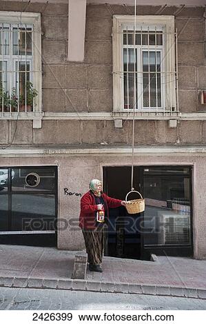 Stock Photo - A woman stands holding a basket which hangs from an upper level of a building; Istanbul, Turkey. Fotosearch