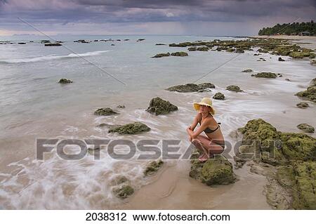 Stock Image - A Woman Tourist Crouches On A Rock On The Beach On A Tropical Island; Koh Lanta Thailand. Fotosearch