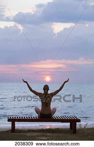 Stock Photo - A Woman Tourist Sits On A Bench With Arms Raised At Sunset On The Beach Of A Tropical Island; Koh Lanta Thailand. Fotosearch