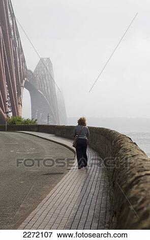 A Woman Walking On A Pathway On The Water's Edge Towards Forth Bridge; North Queensferry Scotland View Large Photo Image Stock Photo - A Woman Walking On A Pathway On The Water's Edge Towards Forth Bridge; North Queensferry Scotland. Fotosearch
