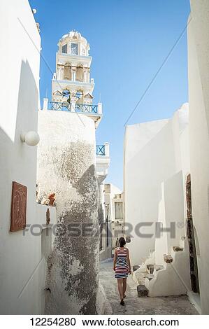 Stock Image - A woman walks between whitewash buildings; Megalochori, Santorini, Greece. Fotosearch