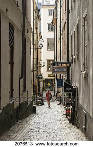 Stock Image - A Woman Walks Down A Narrow Street Between Buildings In Old Town; Stockholm Sweden. Fotosearch