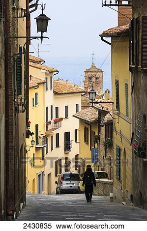 Stock Photography - A woman walks down a narrow street between buildings; Montepulciano, Tuscany, Italy. Fotosearch