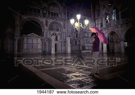 Stock Photo - A Woman Walks Holding A Red Umbrella Under An Illuminated Street Light; Venice Venezia Italy. Fotosearch