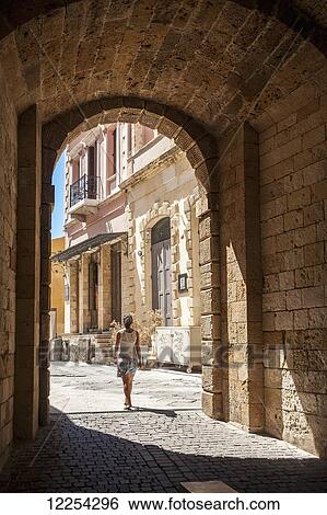 Stock Photograph - A woman walks through an arched walkway; Chania, Crete, Greece. Fotosearch