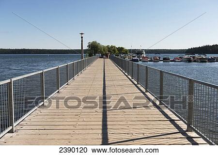 Stock Image - A wooden boardwalk leading out to the water and boats mooring in a harbour; Manitoba, Canada. Fotosearch