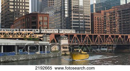 Stock Photography - A Yellow Boat In The River Going Under A Bridge With The Train And Buildings In The Background; Chicago Illinois United States Of America. Fotosearch
