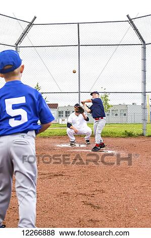 A young boy in team baseball uniform throws the ball to the batter on his team with dad playing back catcher in the baseball diamond; Fort McMurray, Alberta, Canada View Large Photo Image Stock Photo - A young boy in team baseball uniform throws the ball to the batter on his team with dad playing back catcher in the baseball diamond; Fort McMurray, Alberta, Canada. Fotosearch
