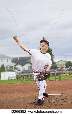 Stock Image - A young boy pitches the baseball from the pitchers mound during a ball game in a white uniform with a basball and glove; Fort McMurray, Alberta, Canada. Fotosearch