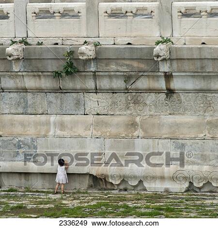 Stock Photo - A Young Girl Standing Against An Old Stone Wall With Gargoyles Along The Top; Beijing, China. Fotosearch