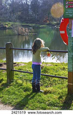 Stock Photo - A young girl stands at a fence looking out over a tranquil lake, Hamstead Heath; London, England. Fotosearch