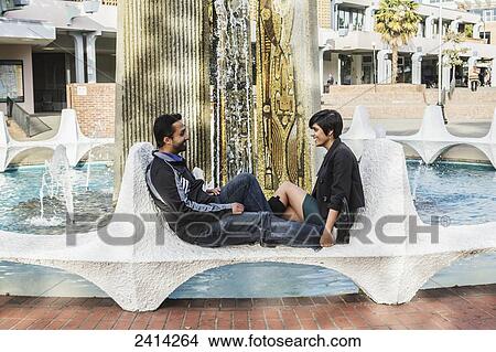 Picture - A young Indian ethnicity couple relaxing at the Ceramic Fins of Centennial Square fountain; Victoria, Vancouver Island, British Columbia. Fotosearch