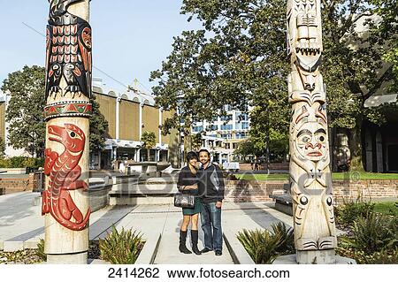 Stock Image - A young Indian ethnicity couple poses by totem poles at the Signs of Lekwungen interpretive walkway; Victoria, Vancouver Island, British Columbia. Fotosearch