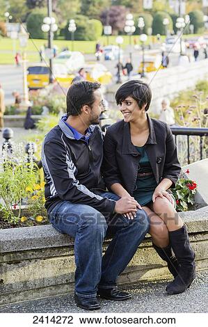 Stock Image - A young Indian ethnicity couple sit together smiling romantically; Victoria, Vancouver Island, British Columbia. Fotosearch