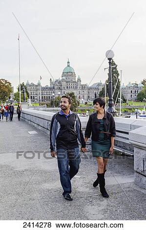Stock Photo - A young Indian ethnicity couple walking the waterfront away from the Parliament Buildings; Victoria, Vancouver Island, British Columbia. Fotosearch