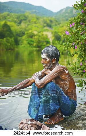 Picture - A young man crouched at the water's edge covered in soap suds; Ulpotha, Embogama, Sri Lanka. Fotosearch