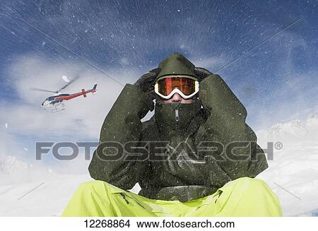 A young man sits in winter apparel and a ski mask while a helicopter flies overhead and blows snow; Methven, New Zealand View Large Photo Image Picture - A young man sits in winter apparel and a ski mask while a helicopter flies overhead and blows snow; Methven, New Zealand. Fotosearch
