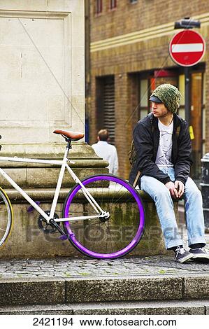 Picture - A young man sits on a ledge beside a bicycle; London, England. Fotosearch