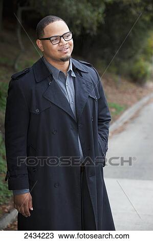 Stock Image - A young man standing on the street wearing a dark trench coat; San Francisco, California, USA. Fotosearch