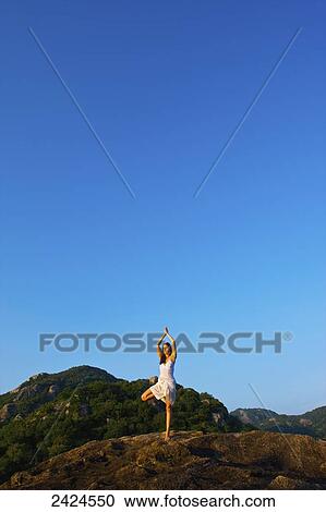 Stock Image - A young woman does a yoga pose on a hilltop against a blue sky; Ulpotha, Embogama, Sri Lanka. Fotosearch