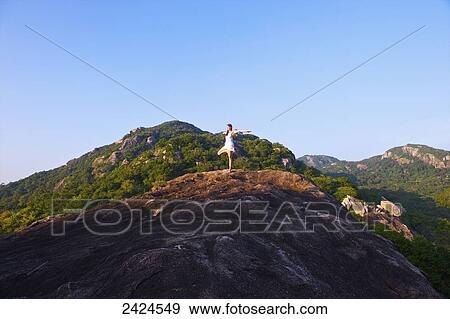 Stock Photo - A young woman does a yoga pose on a hilltop against a blue sky; Ulpotha, Embogama, Sri Lanka. Fotosearch