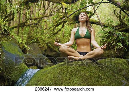 Stock Photo - A young woman in a bikini does a yoga pose on a moss covered rock; Hawaii, United States of America. Fotosearch