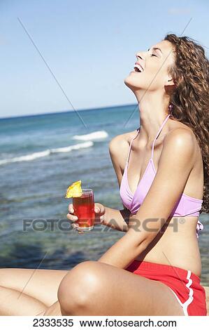A young woman in a bikini holds a drink and laughs while sitting at the water's edge; Honolulu, Oahu, Hawaii, United States of America View Large Photo Image Stock Photography - A young woman in a bikini holds a drink and laughs while sitting at the water's edge; Honolulu, Oahu, Hawaii, United States of America. Fotosearch