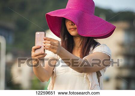 Stock Image - A young woman in a large brimmed pink hat takes a self-portrait with her cell phone; Xiamen, Fujian, China. Fotosearch