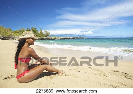 Stock Photo - A young woman in a red bikini sitting on the beach of an hawaiian island; Maui, Hawaii, United States of America. Fotosearch