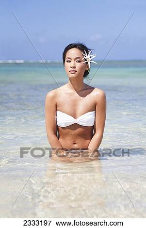 Stock Photo - A young woman in a white bikini sits in the shallow ocean water with Tiger Lilies in her hair; Oahu, Hawaii, United States of America. Fotosearch