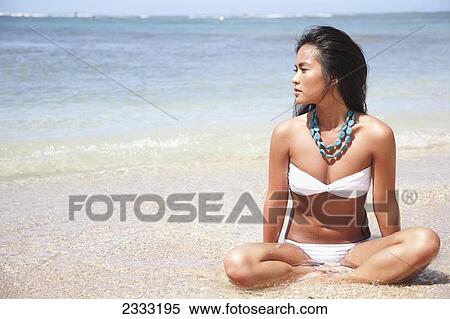 Stock Photography - A young woman in a white bikini sits in the shallow ocean water; Oahu, Hawaii, United States of America. Fotosearch
