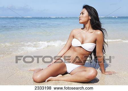 A young woman in a white bikini sits on the beach by the ocean water; Oahu, Hawaii, United States of America View Large Photo Image Stock Photo - A young woman in a white bikini sits on the beach by the ocean water; Oahu, Hawaii, United States of America. Fotosearch