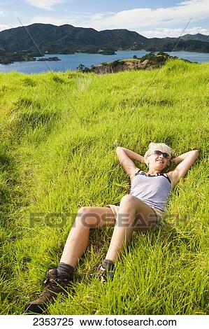 Stock Photography - A Young Woman Lays On The Grass With A View Of The Coastline; Urupukapuka Island, New Zealand. Fotosearch