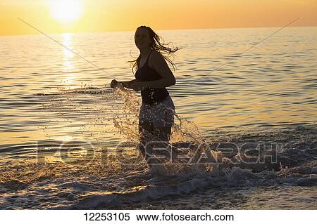 Stock Photography - A young woman running in the shallow water at sunset; Nokomis, Florida, United States of America. Fotosearch