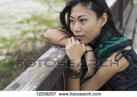 Stock Photo - A young woman sits against a wooden railing looking out at the ocean; Jincheng, Kinmen, Taiwan. Fotosearch