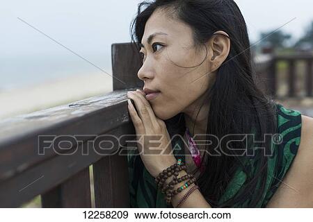 Stock Photo - A young woman sits against a wooden railing looking out at the ocean; Jincheng, Kinmen, Taiwan. Fotosearch