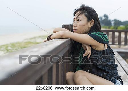A young woman sits against a wooden railing looking out at the ocean; Jincheng, Kinmen, Taiwan View Large Photo Image Stock Photograph - A young woman sits against a wooden railing looking out at the ocean; Jincheng, Kinmen, Taiwan. Fotosearch