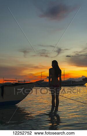 Stock Image - A young woman stands in the shallow water beside a boat at Alona beach at sunset; Panglao Island, Bohol, Philippines. Fotosearch