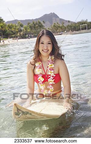 Stock Photo - A young woman stands in the water wearing a flower lei with a surfboard; Honolulu, Oahu, Hawaii, United States of America. Fotosearch