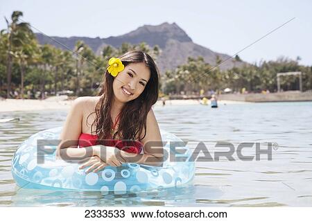 Stock Image - A young woman with a tropical flower in her hair standing in the water with an inflatable tube; Honolulu, Oahu, Hawaii, United States of America. Fotosearch