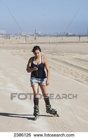 Stock Photograph - A young woman with inline skates on the promenade along the coast with earbuds and a cell phone; California, United States of America. Fotosearch