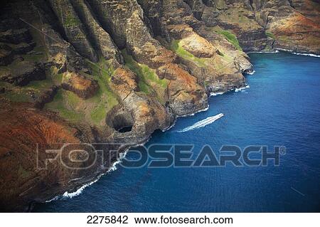 Stock Image - Aerial view of the rugged coastline and a boat in the pacific ocean along an hawaiian island; Na Pali coast of Kauai, Hawaii, United States of America. Fotosearch