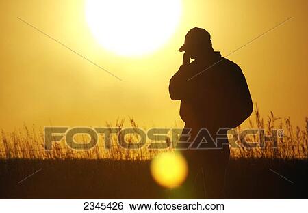 Stock Photograph - Agriculture - A farmer, silhouetted by the setting sun, talks on a cell phone in his field / Iowa, USA.. Fotosearch