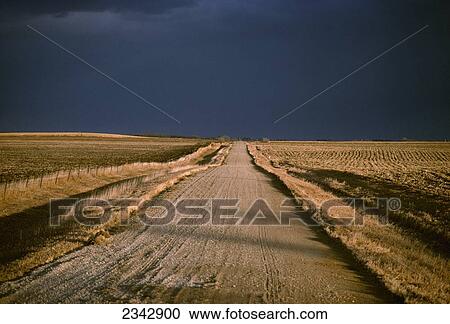 Stock Image - Agriculture - Gravel country road passing through harvested farm fields in Autumn with dark storm clouds above / Iowa, USA.. Fotosearch