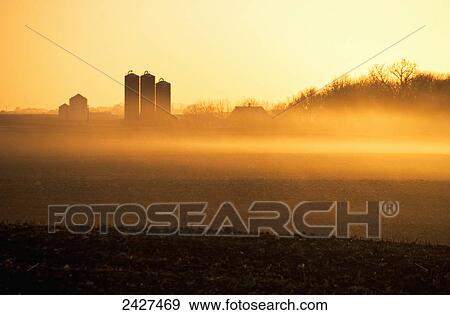 Stock Photo - Agriculture - Hazy sunrise light on fallow cornfields and a farmstead with Harvestore silos / near Truman Minnesota, USA.. Fotosearch