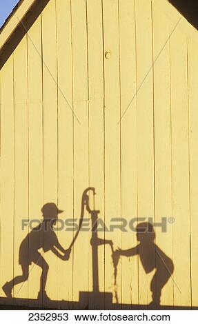 Stock Image - Agriculture - Silhouette on the side of a barn in late afternoon light of a farm boy operating a hand pump water well while his young sister washes her hands / Northwest Missouri, USA.. Fotosearch
